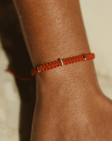 A close up of a model's wrist showcasing a red woven thread bracelet with three 14k Yellow Gold tabs evenly spaced out, with one tab having 5 diamonds on it.