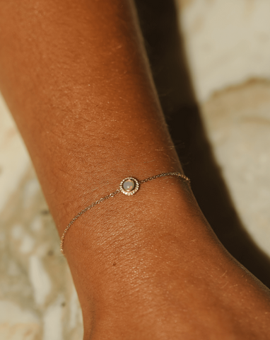 A close up shot of a 14k Yellow Gold delicate bracelet featuring a small opal stone encircled by tiny white sapphires, elegantly displayed on the model's wrist.