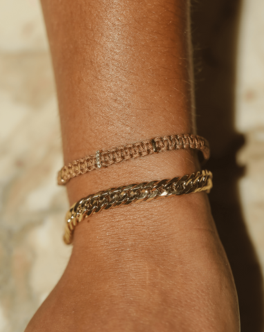 A close up of a model's wrist showcasing a tan woven thread bracelet with three 14k Yellow Gold tabs evenly spaced out, with one tab having 5 diamonds on it, paired with a thick curb chain bracelet. The model’s wrist is laid against a marble surface.
