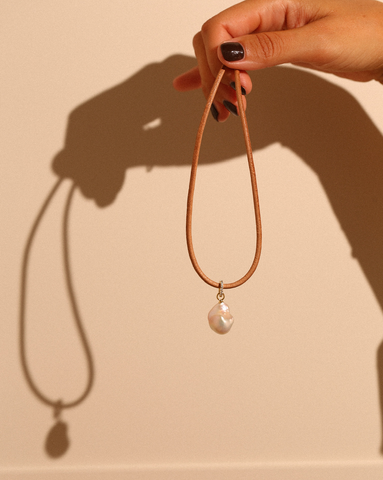 Model's hand is holding a leather necklace with a fresh water pearl charm attached with a gold vermeil pave charm clasp.